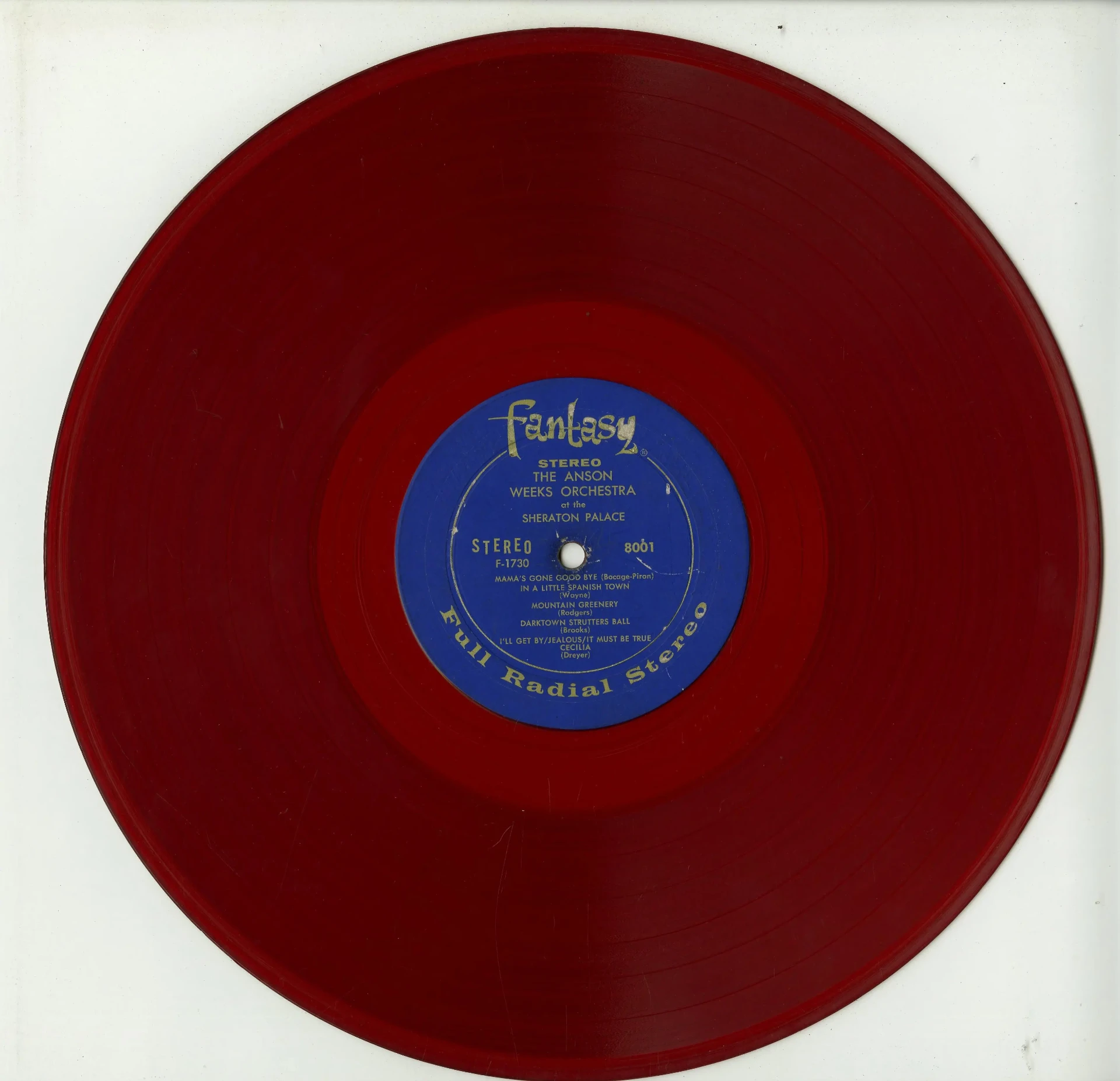 Vintage translucent ruby-red vinyl record “The Anson Weeks Orchestra at the Sheraton Palace (1900)” with a blue center label, photographed against white; nostalgic, collectible music memorabilia.