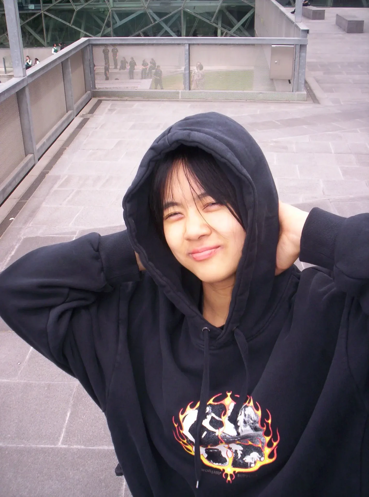 Person in a black hoodie with a flame design stands outdoors on gray steps, smiling at the camera with hands behind their head.